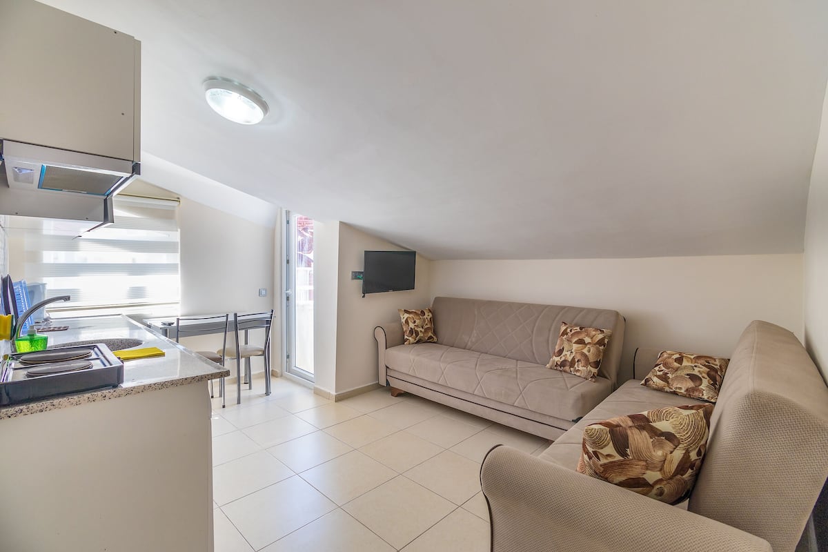 A modern kitchen with a built-in stove, a dining area with chairs, a sofa with patterned cushions, and a flat-screen TV mounted on the wall.