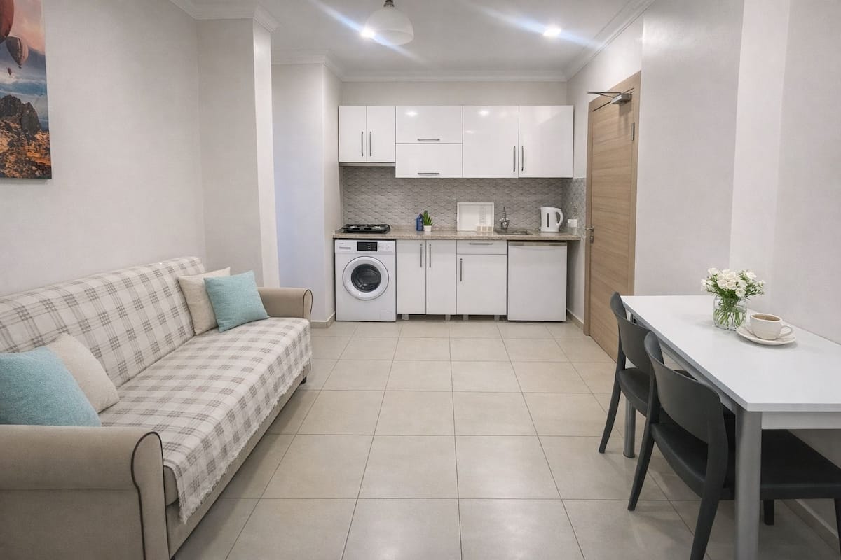 A compact living space with a sofa, a dining table, a washing machine, and a kitchenette.