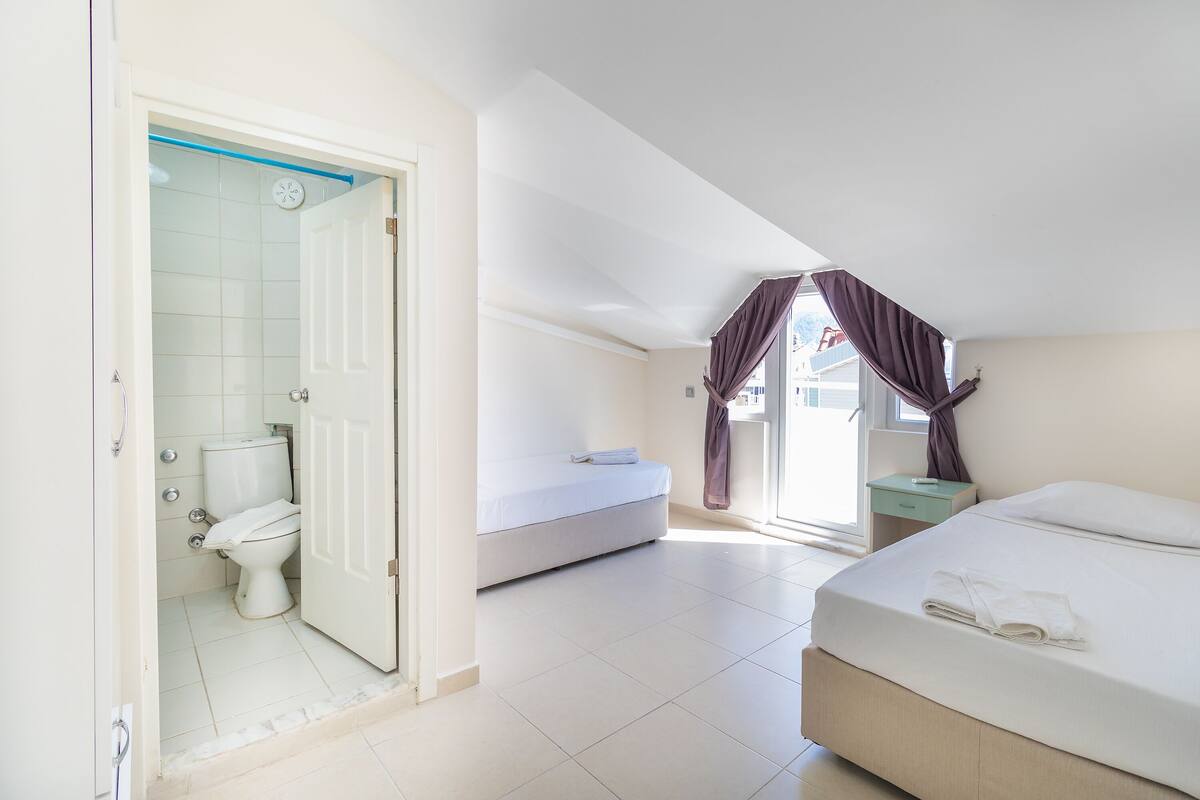 A hotel room with a bed, a bedside table, a window with curtains, and a bathroom with a toilet and sink.