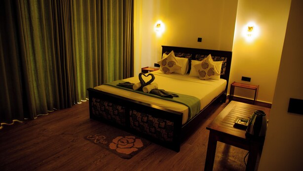 A neatly made bed with a heart-shaped pillow, a bedside table with a kettle, and a small nightstand with a lamp. - Jade Ella (Ella)