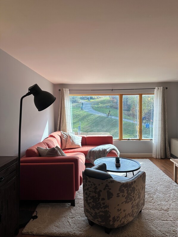 A living room with a red sofa, a round ottoman, a floor lamp, and a large window with a view of a green landscape.