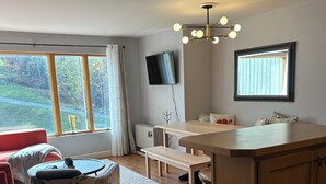 A modern living room with a large window, a flat-screen TV, a wooden dining table, and a cozy seating area with a red sofa. - One Bedroom Ski-In/Ski-Out Condo on Whiffletree Chairlift. Short Walk to Village (Carrabassett Valley)