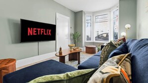 Una sala de estar con una televisión de pantalla plana mostrando 'NETFLIX', un sofá, una mesa de centro y una ventana con persianas.
