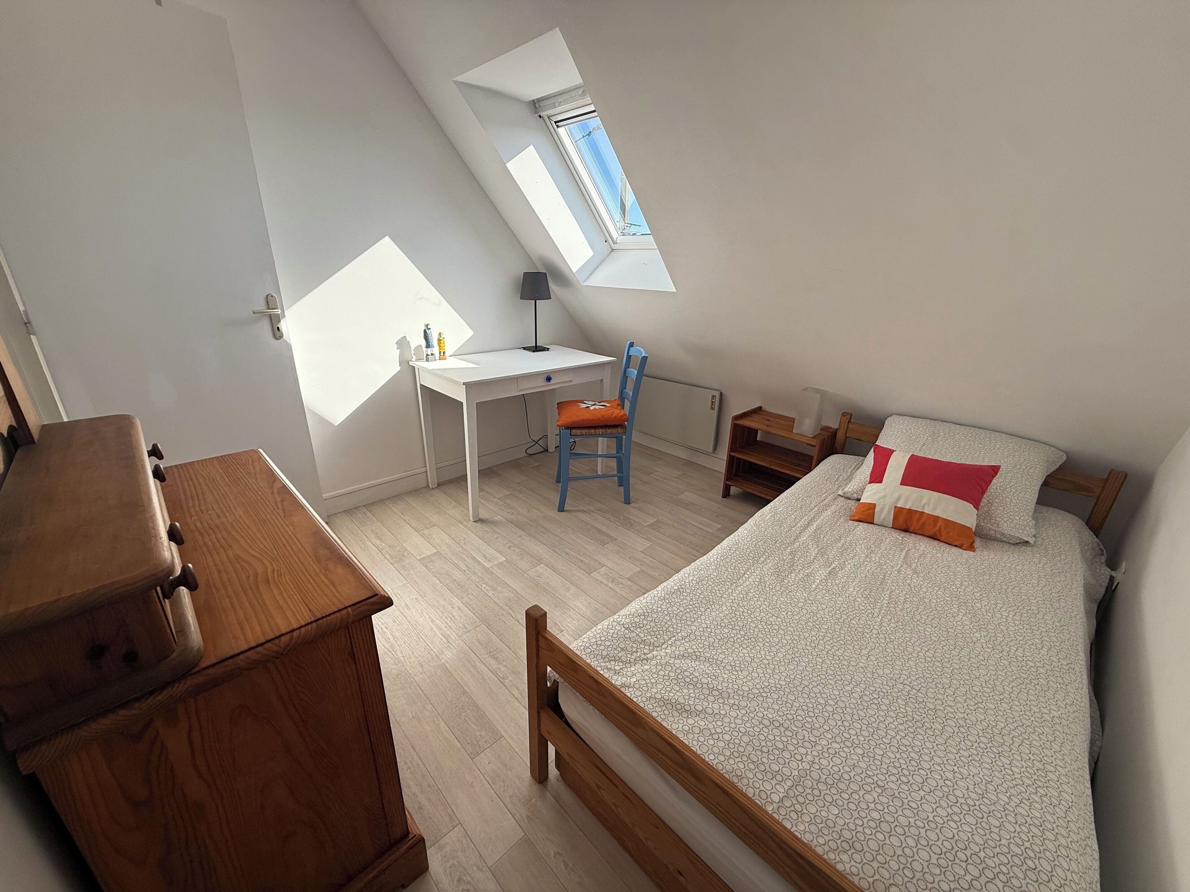 Une chambre avec un plafond en pente, un bureau, une chaise, un lit avec une literie à motifs et une commode.