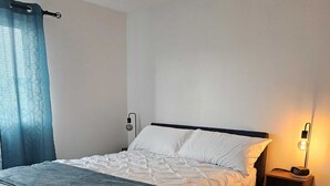 Una cama bien hecha con un cabecero oscuro, una mesita de noche con lámpara y una cortina azul entreabierta.