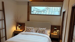 Une chambre dotée d’un grand lit, de poutres apparentes, d’une lucarne et offrant une vue sur une architecture traditionnelle.