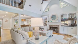 A spacious living room with a high ceiling, a ceiling fan, a fireplace, and a large window.