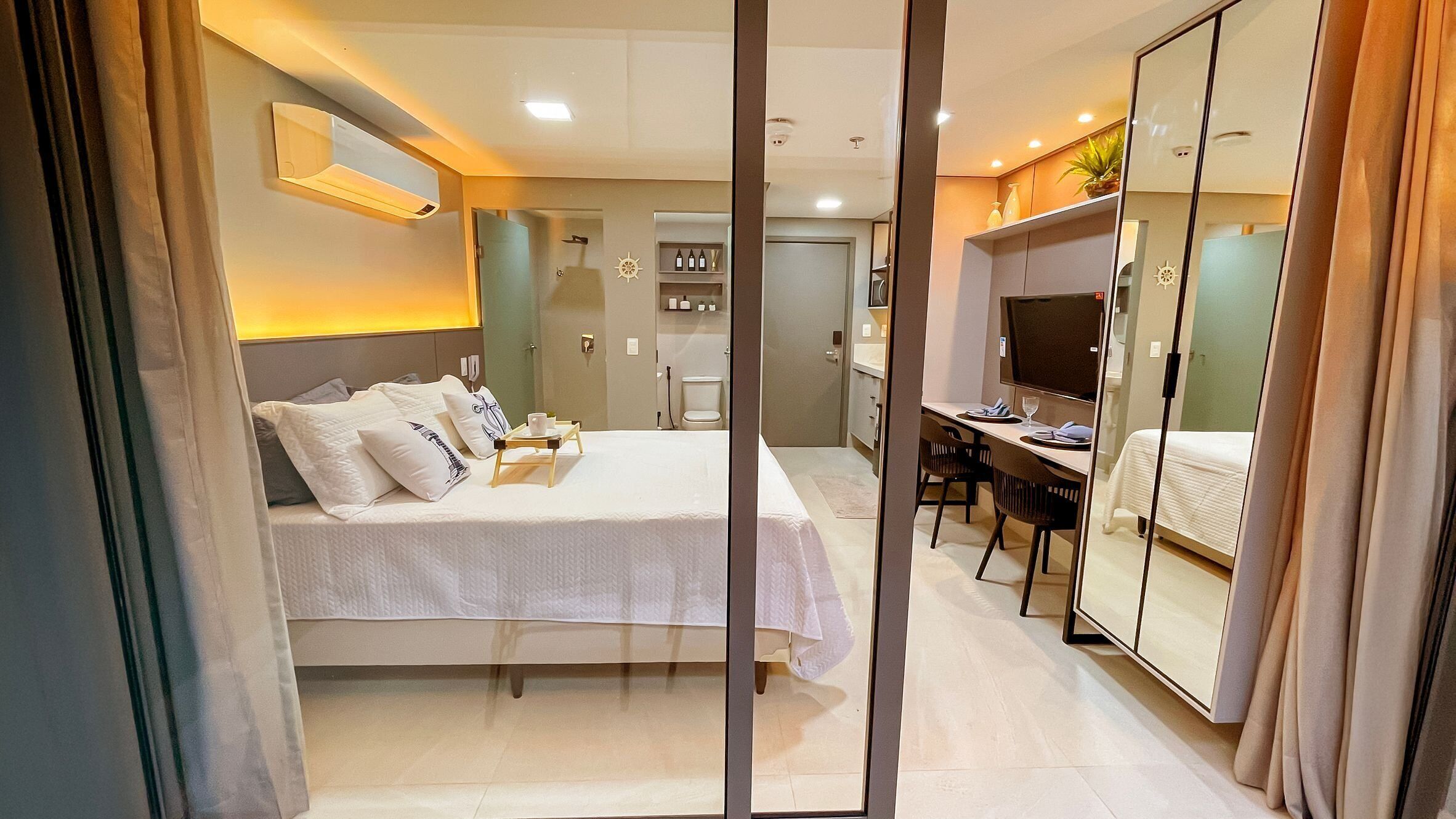 A modern hotel room with a bed, bedside table, mirror, desk, and a TV.