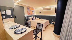 Een compacte hotelkamer met een bed, een klein tafeltje gedekt voor twee personen, een stoel, een wandairconditioner en een groot raam.