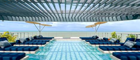 A rooftop infinity pool with blue lounge chairs and umbrellas, overlooking a beach.