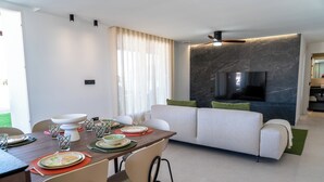 A modern living room with a dining area, a flat-screen TV mounted on a marble wall, and a sofa with green cushions. - Luxury Apartment in La Zenia (Orihuela)