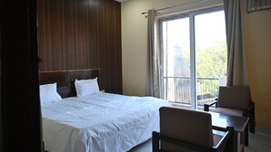 Una habitación de hotel con una cama grande, cabecera de madera, balcón con una silla y ventana con cortinas.