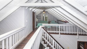 A high-ceilinged attic bedroom with white railings, a wooden staircase, and a bed with a green bedspread.