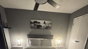 Ein Schlafzimmer mit einem Bett, Deckenventilator, Nachttischlampen, einem Nachttisch, einer Tür und einem gerahmten Bild an der Wand.