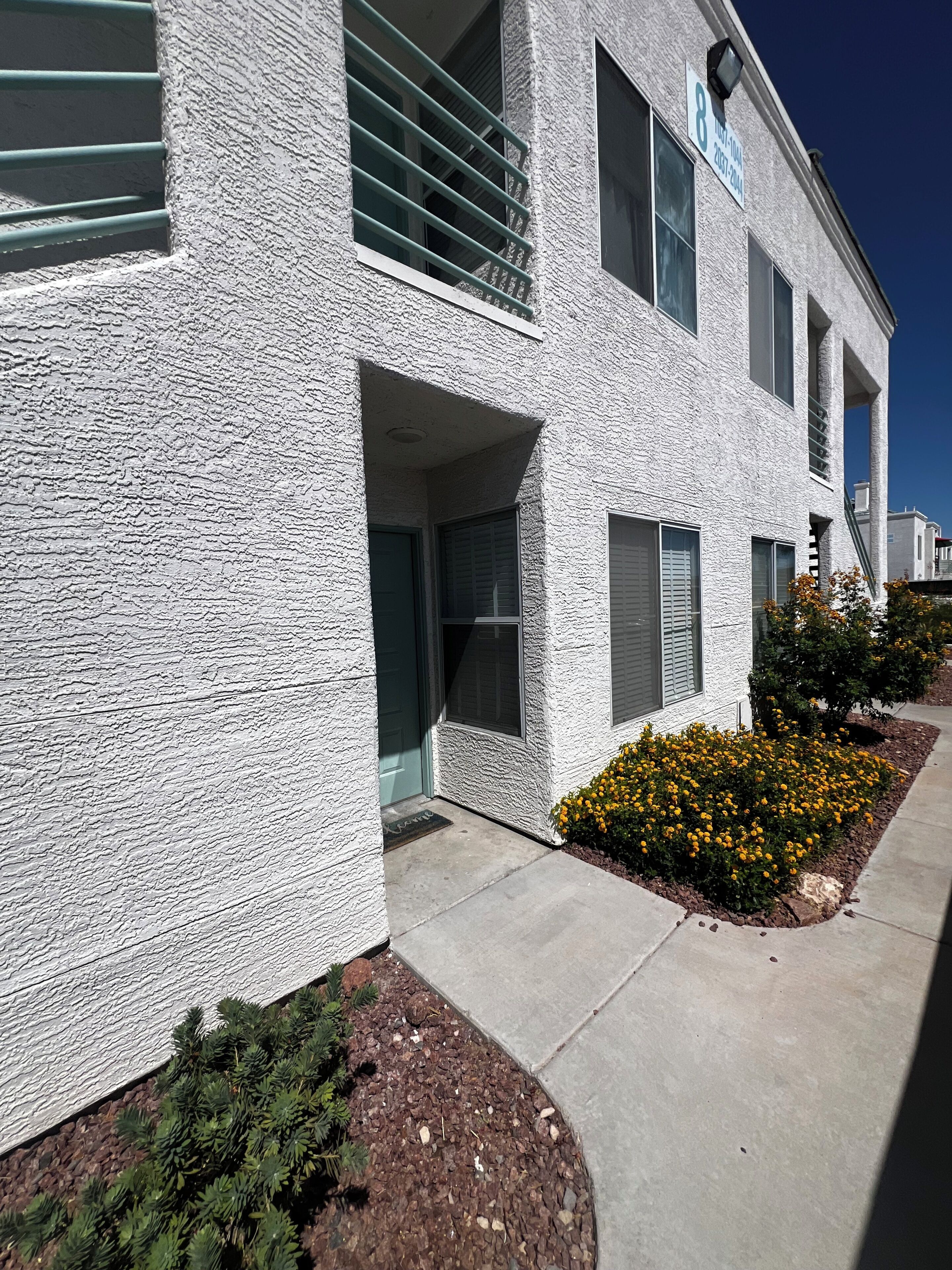 A building with a textured facade, multiple windows, and a small garden with yellow flowers.