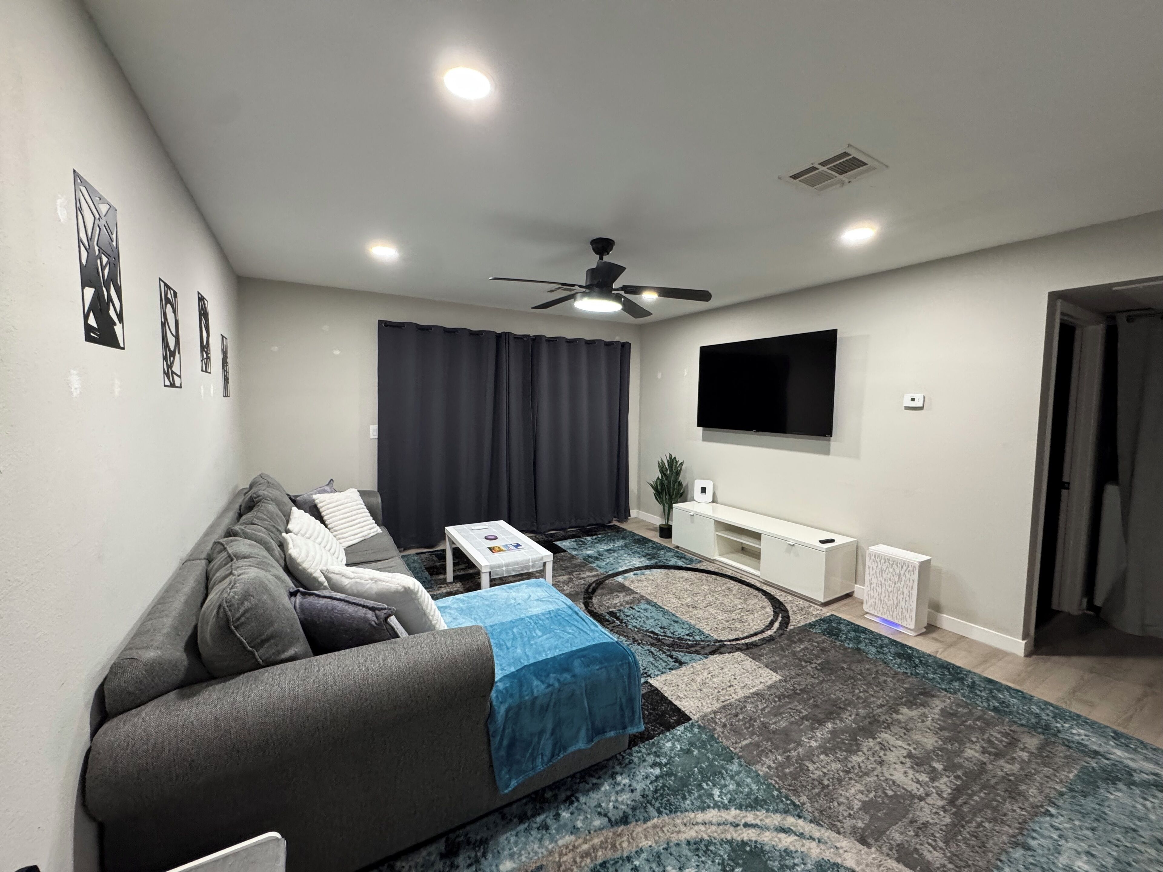 Una sala de estar moderna con un sofá gris, una mesa de centro, un televisor montado en la pared y un ventilador de techo.