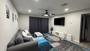 A modern living room with a grey sofa, a coffee table, a TV mounted on the wall, and a ceiling fan. - Zen Den with Pool! (Las Vegas)