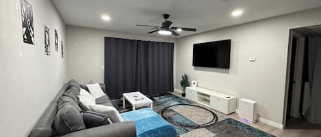 Una sala de estar moderna con un sofá gris, una mesa de centro, un televisor montado en la pared y un ventilador de techo.