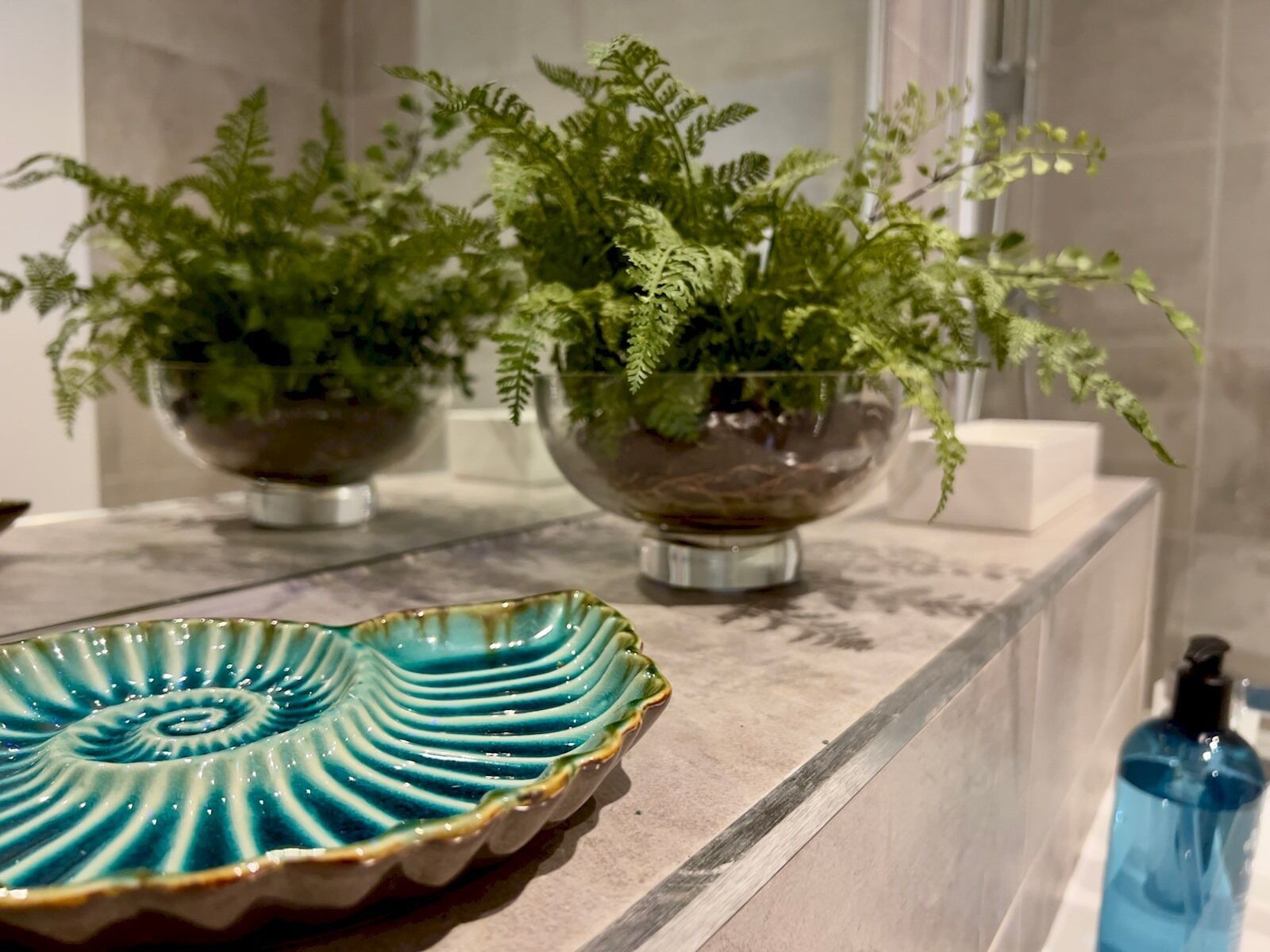 Un bagno con un piano di marmo, due ciotole di vetro con piante, un piatto di ceramica blu e una bottiglia di liquido blu.