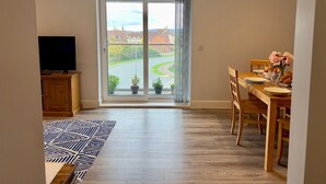 A spacious room with a large sliding glass door leading to a view of a road and houses. There is a dining table with chairs, a television on a wooden cabinet, and a patterned rug on the floor. - Pass the Keys |Modern Penthouse Style Apartment in Haddenham (Aylesbury)