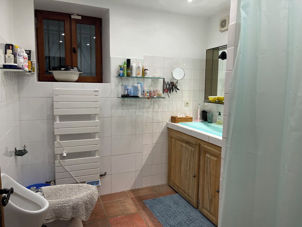 Une salle de bain avec un mur recouvert de carreaux blancs, un meuble en bois, un miroir et une fenêtre avec un pot de fleurs posé sur le rebord.