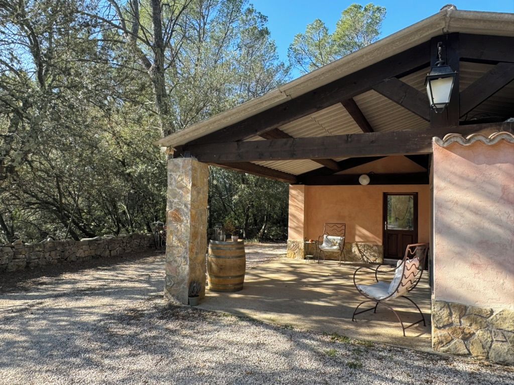 A covered outdoor seating area with stone pillars, a wooden pergola, and a rustic building with a door and windows.