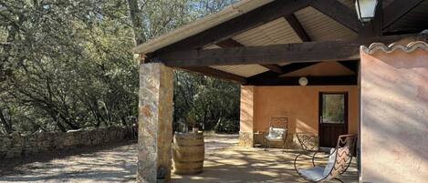 A covered outdoor seating area with stone pillars, a wooden pergola, and a rustic building with a door and windows.