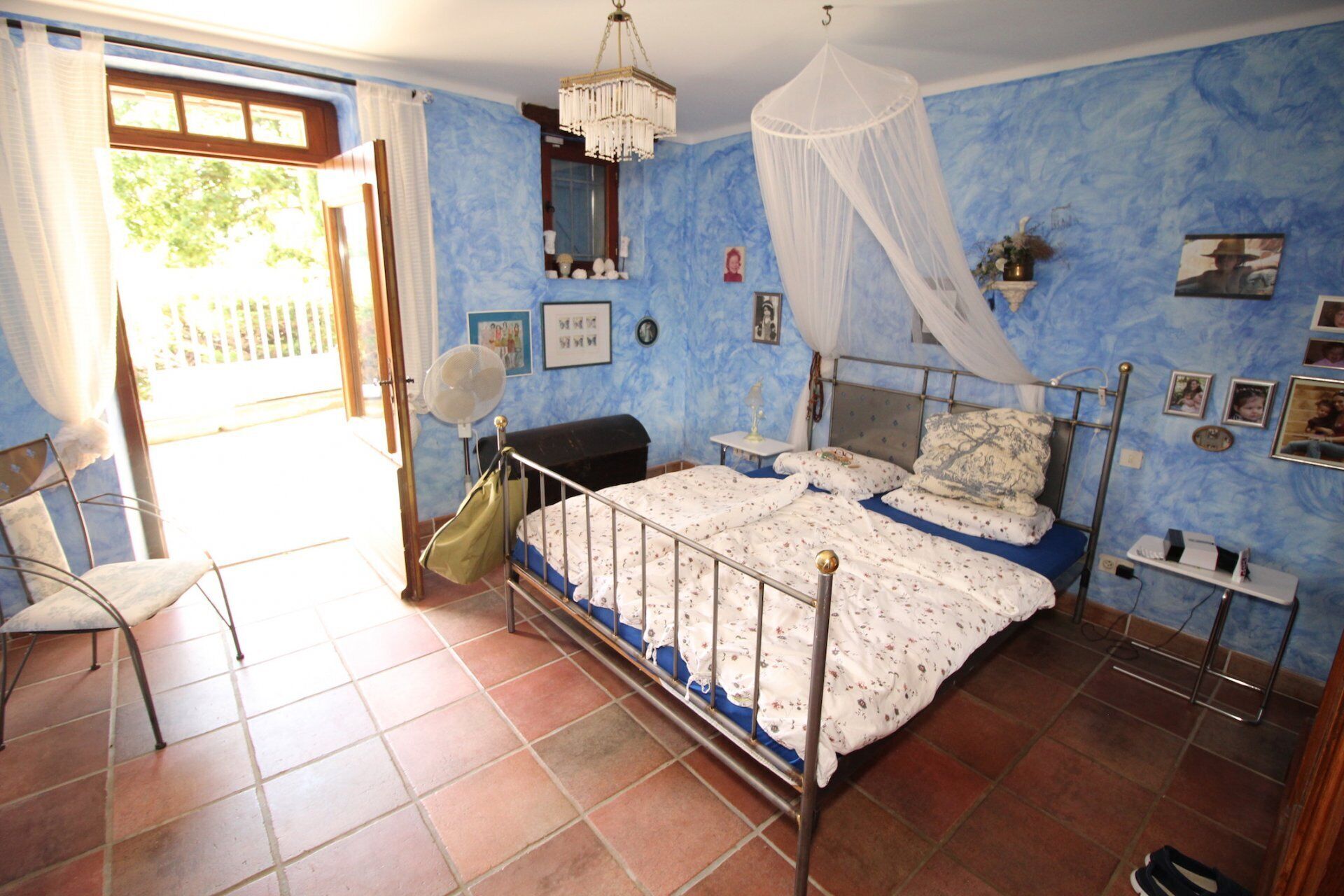 Ein Schlafzimmer mit Bett, Sessel, Ventilator und einem Fenster mit Vorhängen.