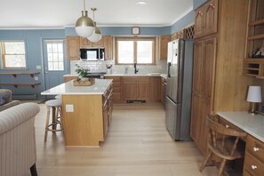 A kitchen with wooden cabinets, a stainless steel refrigerator, a central island, and a hanging light fixture. - Grand Isle Lakehouse with Hot Tub (Grand Isle)