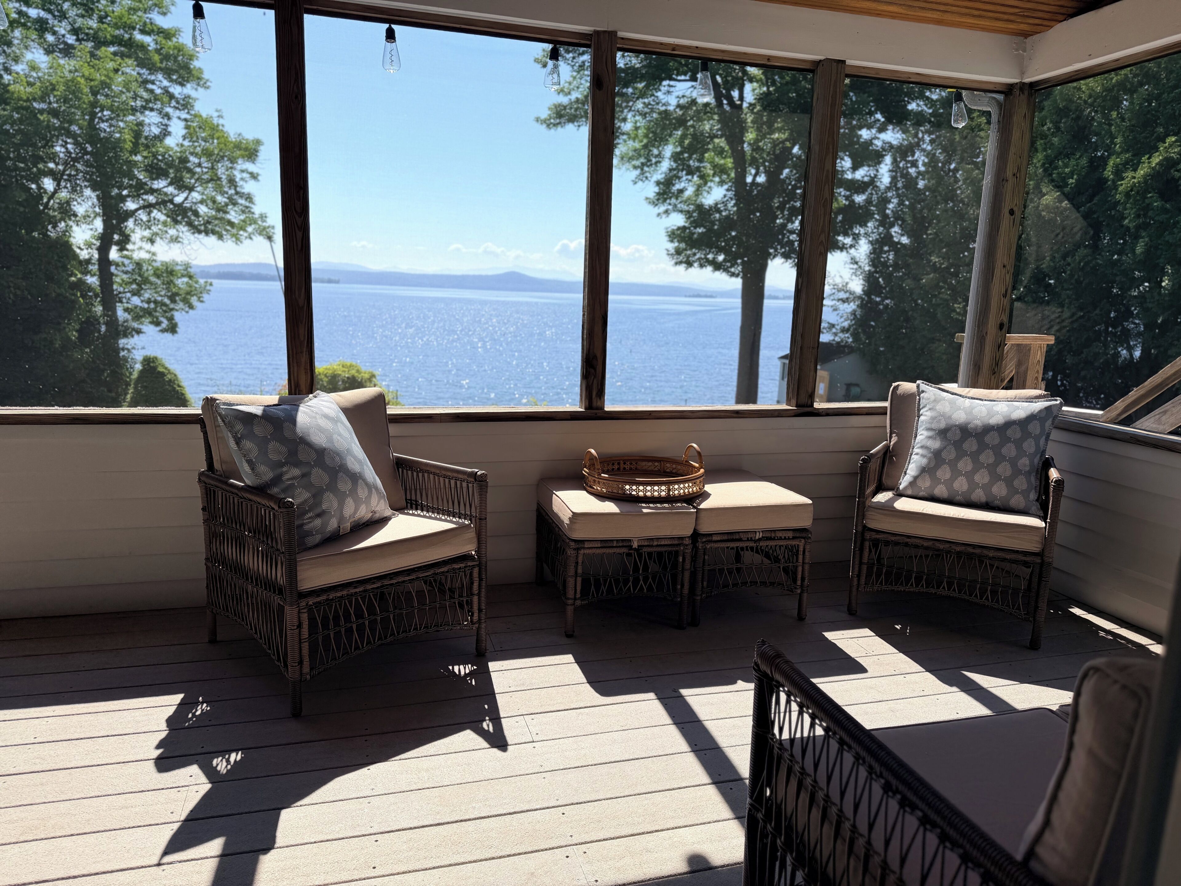 A sunlit room with wicker furniture, a small table, and a view of a body of water through large windows.