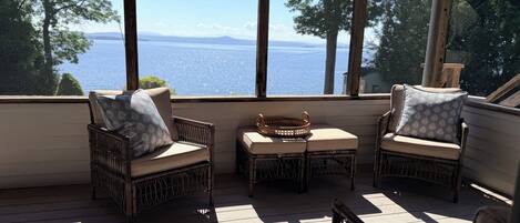 A sunlit room with wicker furniture, a small table, and a view of a body of water through large windows.