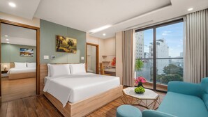 A modern hotel room with a large bed, a seating area with a blue sofa, a small round table with a vase of flowers, and a large window offering a city view. - Happy House Hotel & Apartment (Da Nang)