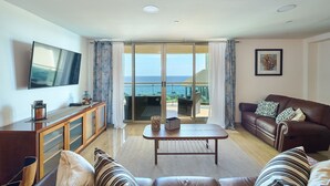 Ein Wohnzimmer mit einer Couch, einem Couchtisch und einem Fernseher. Es gibt einen Balkon mit Meerblick.