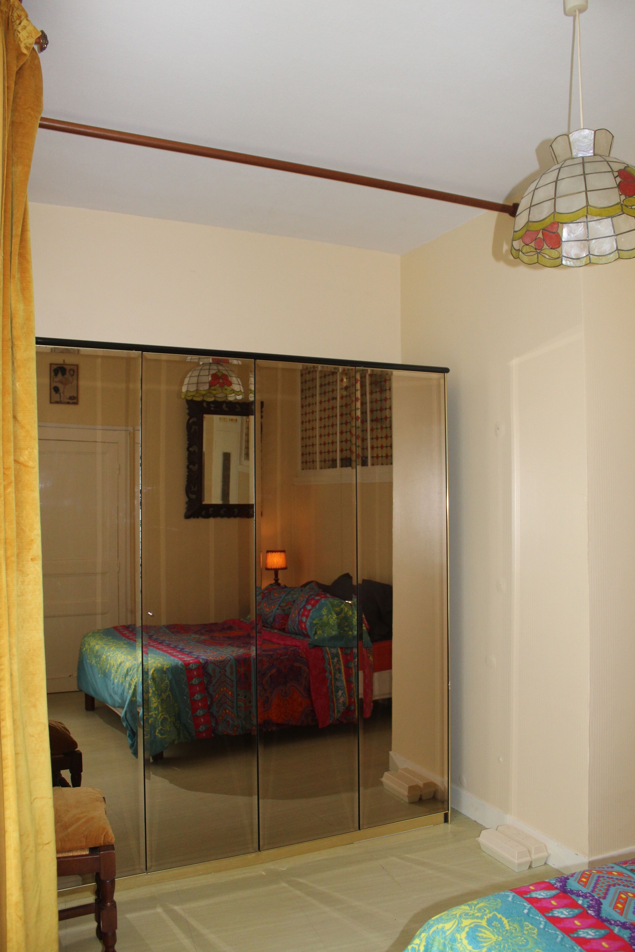 A bedroom with a mirrored wardrobe, a bed with colorful bedding, a bedside table, and a hanging lamp.
