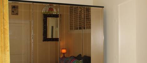 A bedroom with a mirrored wardrobe, a bed with colorful bedding, a bedside table, and a hanging lamp.
