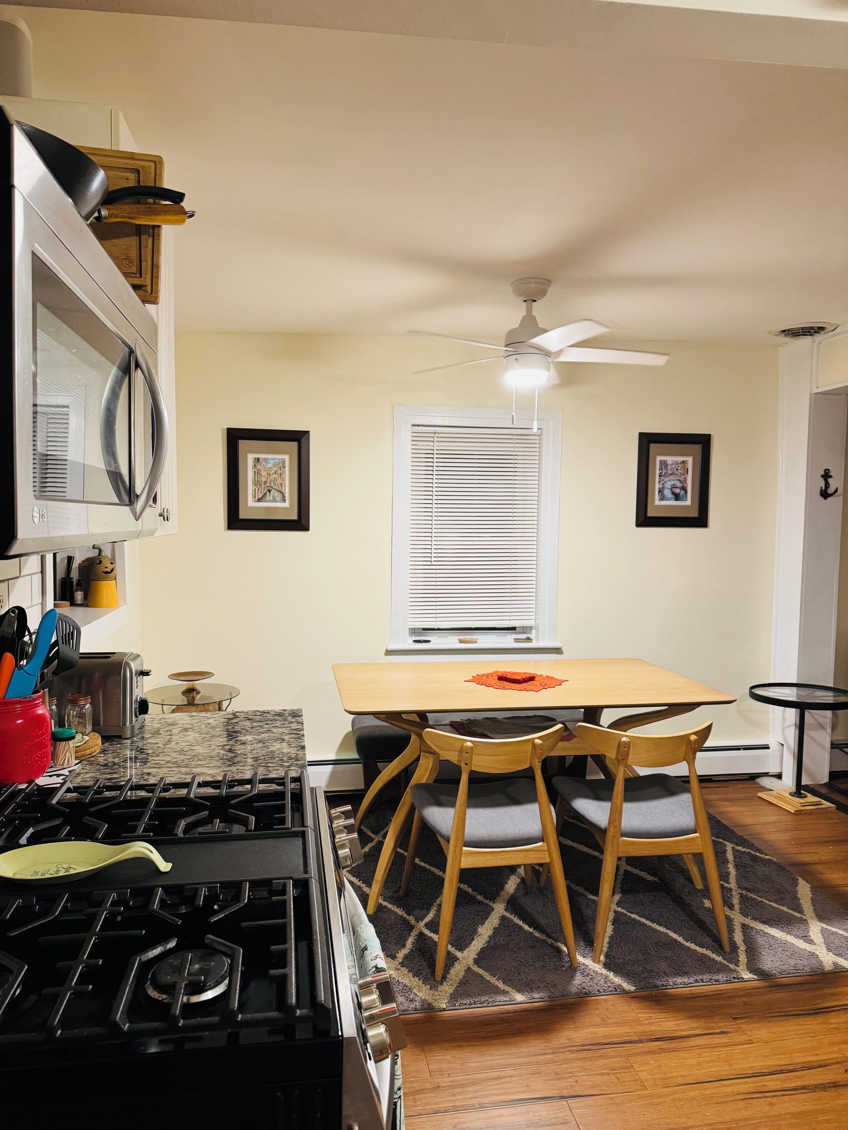 A kitchen with a stove, microwave, and dining table with chairs. There are framed pictures on the wall and a ceiling fan.