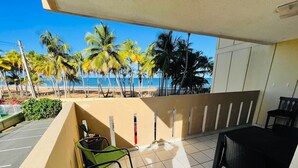 Un balcón con vista a la playa y palmeras, que incluye dos sillas verdes y una mesa negra.