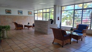 Un vestíbulo amplio con suelo de baldosas, mobiliario de mimbre, un mostrador de recepción y grandes ventanales con vistas a palmeras.