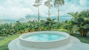 A circular pool with a surrounding stone deck, surrounded by tropical vegetation and tall trees under a partly cloudy sky. - Hideout Kintamani and Hot Spring (Kintamani)