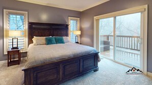 A bedroom with a large bed, two bedside lamps, a dark wooden headboard, and a view of a balcony through a sliding glass door. - Breeze on Inn • Deck, Firepit, & Game Room (Sugar Mountain)