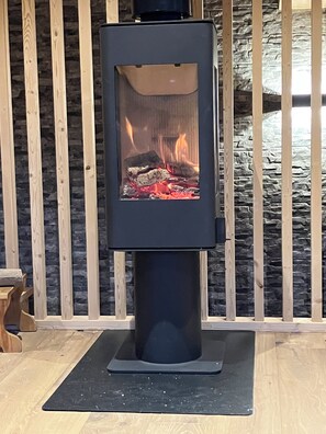 A wood-burning stove with visible logs and flames, set against a backdrop of wooden slats and a stone wall. - New Chalet in Pelvoux-Vallouise (Vallouise-Pelvoux)