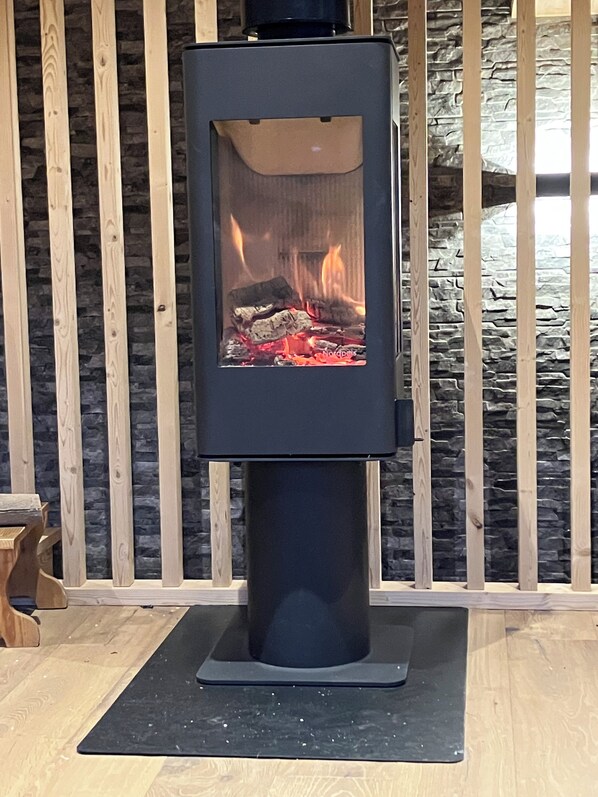 A wood-burning stove with visible logs and flames, set against a backdrop of wooden slats and a stone wall.