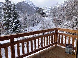 A wooden deck with a metal railing, overlooking a snowy landscape with trees and mountains.