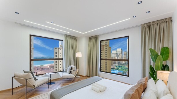 Una habitación de hotel moderna con una cama grande, una zona de estar con una silla y una mesa, y ventanas amplias con vistas a la ciudad.