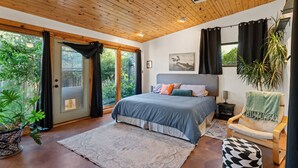 A bedroom with a bed, a chair, a plant, and a door leading to a balcony.