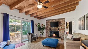 A living room with a fireplace, wooden ceiling beams, a TV mounted on the wall, and a large window with curtains.