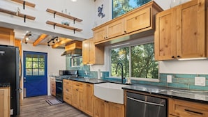 A modern kitchen with wooden cabinets, a black refrigerator, stainless steel dishwasher, and a window with a view of trees.