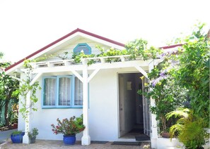 A white house with a red roof, a blue window, and a porch with potted plants.