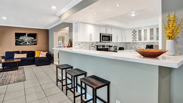A modern kitchen with white cabinetry, a central island, and a dining area with a sofa and bar stools. - Spacious Villa with Heated Pool, Trampoline, BBQ, Tennis, Air Hockey (Hollywood)