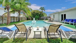 A swimming pool with lounge chairs and a small table, surrounded by palm trees and a fence. - Spacious Villa with Heated Pool, Trampoline, BBQ, Tennis, Air Hockey (Hollywood)